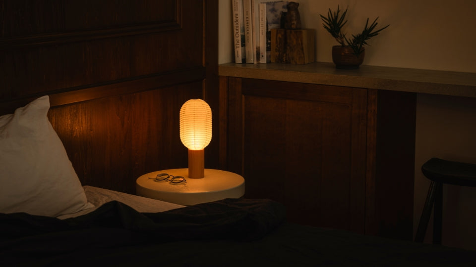 A bedroom with a bed on the left and a bookshelf with books on the right. On the bedside table to the right of the bed is a bedside paper lamp "TORCHIN" made from Japanese lanterns, emitting a warm lamp from its cylindrical shade.