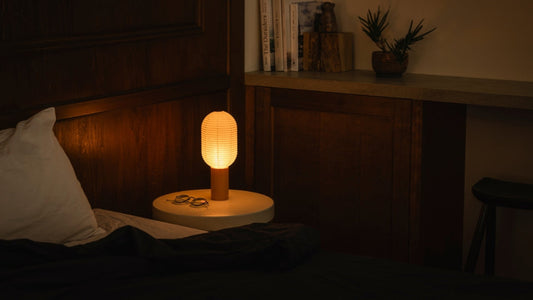 A bedroom with a bed on the left and a bookshelf with books on the right. On the bedside table to the right of the bed is a bedside paper lamp "TORCHIN" made from Japanese lanterns, emitting a warm lamp from its cylindrical shade.