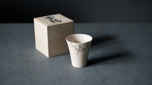 White kintsugi cup with red crackle lines stands next to a wooden box, a meaningful japanese gift representing wabi sabi.