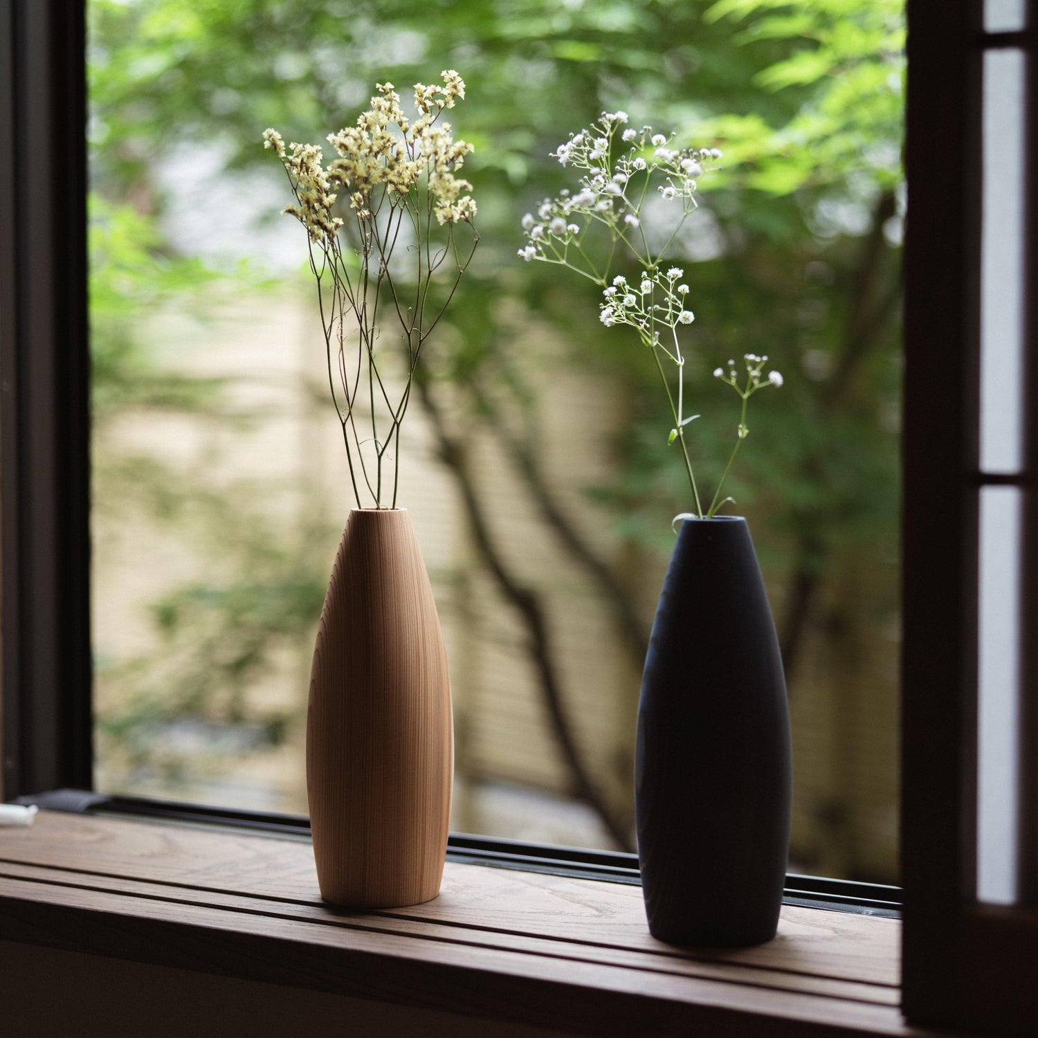 Two minimalist vases crafted from natural Japanese cedar with indigo dye accents, displayed on a windowsill in a Japanese-style bedroom overlooking lush green foliage.