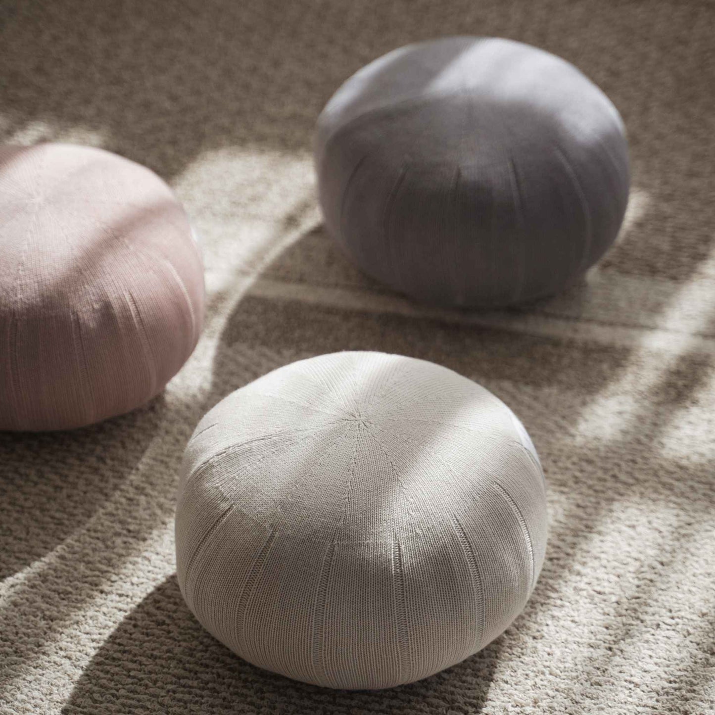 Three zafu meditation cushions in misty rose white gray and lagoon gray arranged on a rug with sunlight shadows.