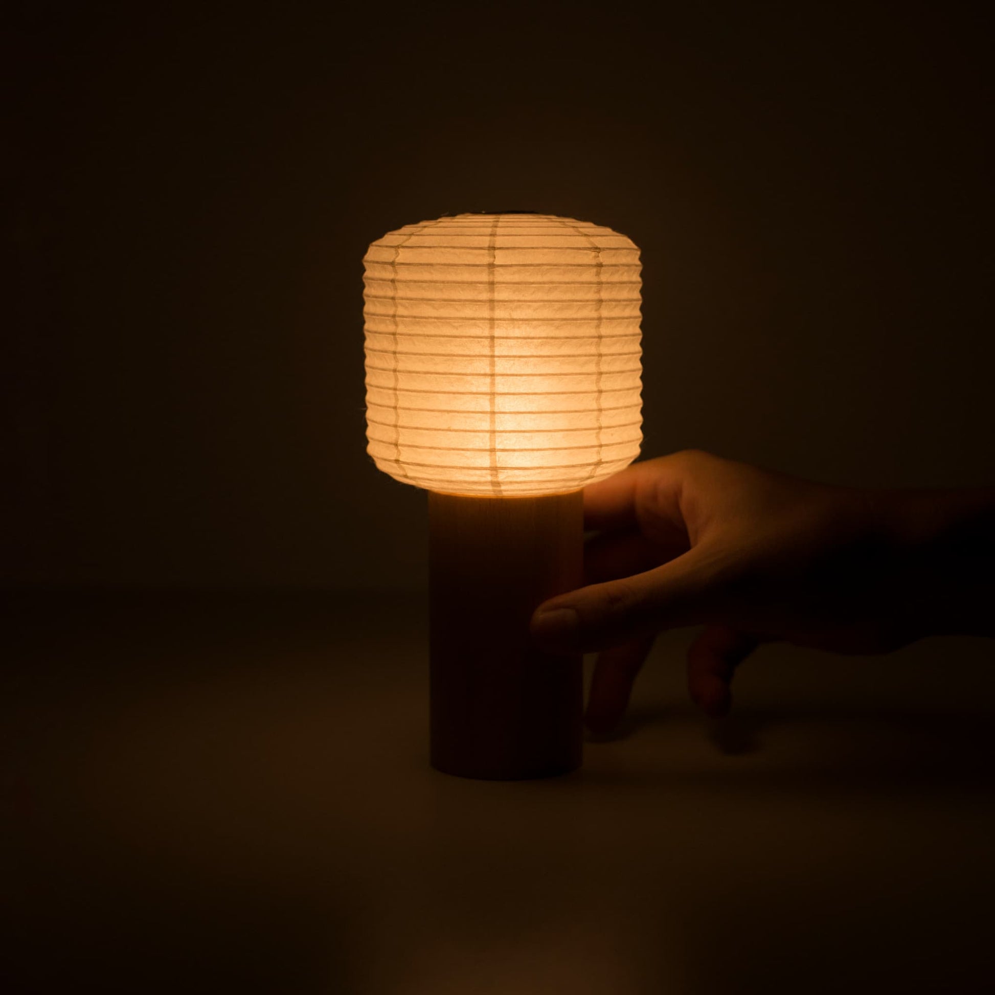 A hand holding a warmly glowing portable paper table lamp in a dark room