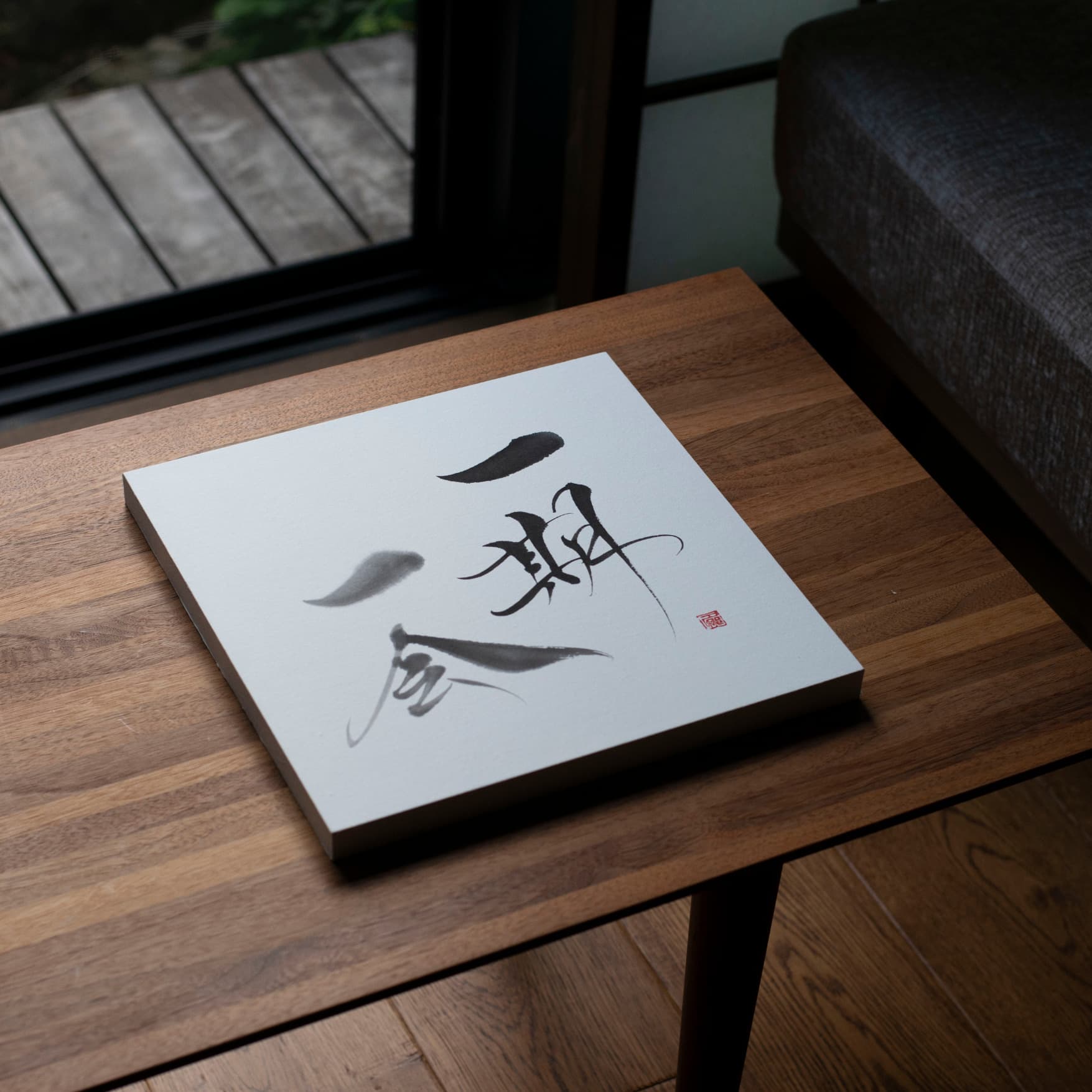 Angled top-down view of a panel with Japanese calligraphy of 'Ichigo Ichie' on a wooden table.