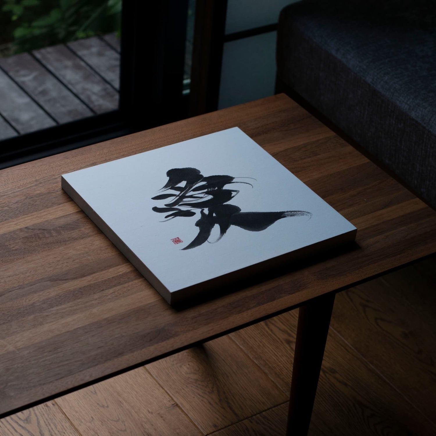 Angled top-down view of a square panel displaying the Japanese calligraphy of '愛' (love) on a wooden table.