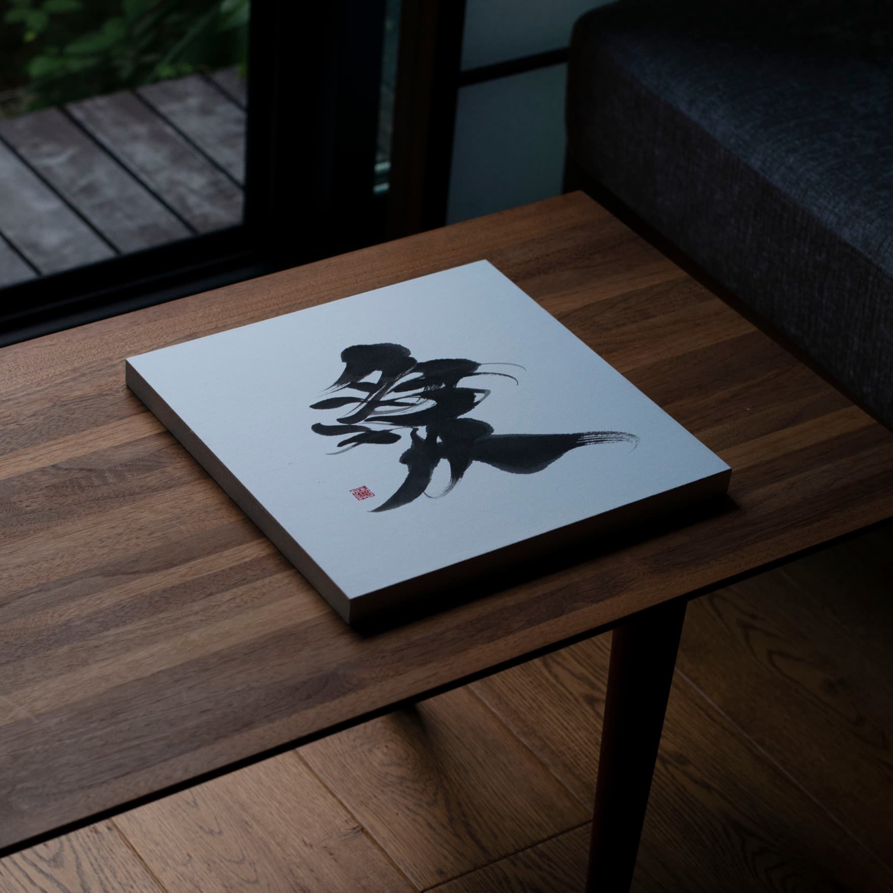 Angled top-down view of a square panel displaying the Japanese calligraphy of '愛' (love) on a wooden table.