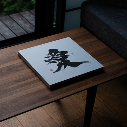 Angled top-down view of a square panel displaying the Japanese calligraphy of '愛' (love) on a wooden table.