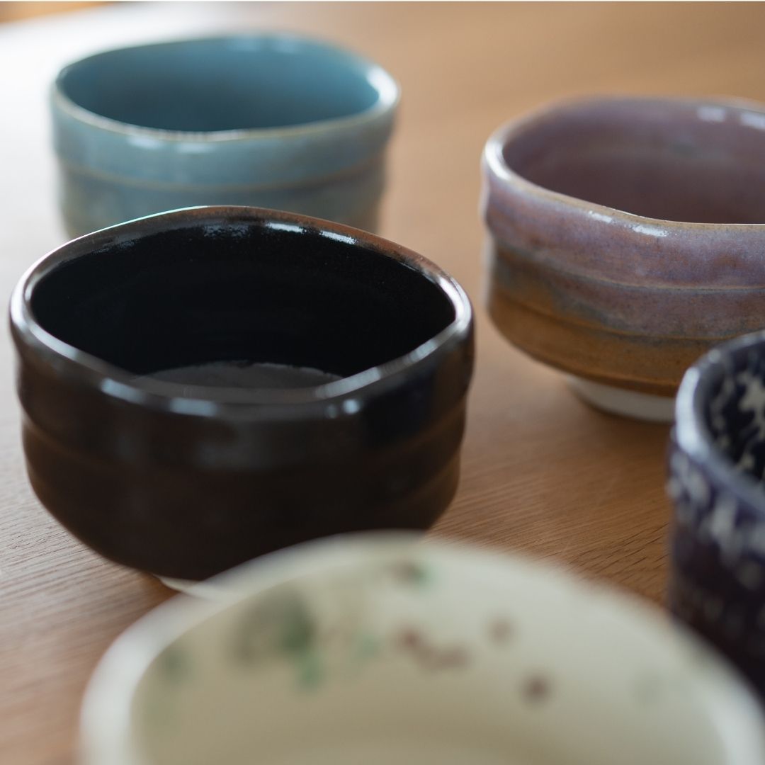 A grouping of five handmade Japanese Mino Ware matcha bowls in various colors including black, blue, and purple, resting on a light wooden table.