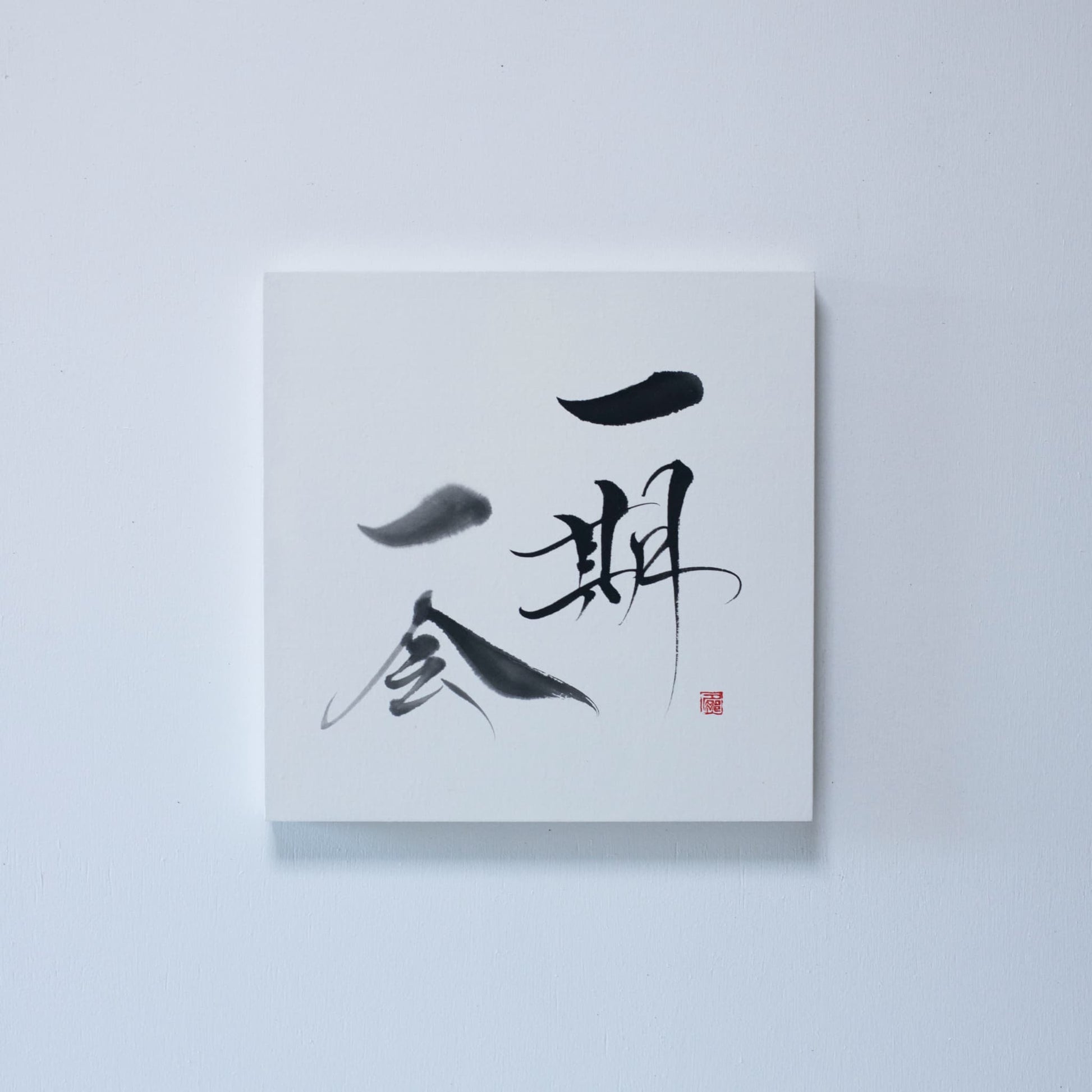 Square panel with traditional Japanese calligraphy of 'Ichigo Ichie' on white rice paper, mounted on a white wall
