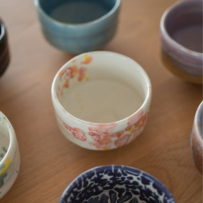 A close-up of a white Mino Ware matcha chawan bowl with delicate pink and orange floral glaze, surrounded by other colorful handmade Japanese matcha bowls on a wooden surface.