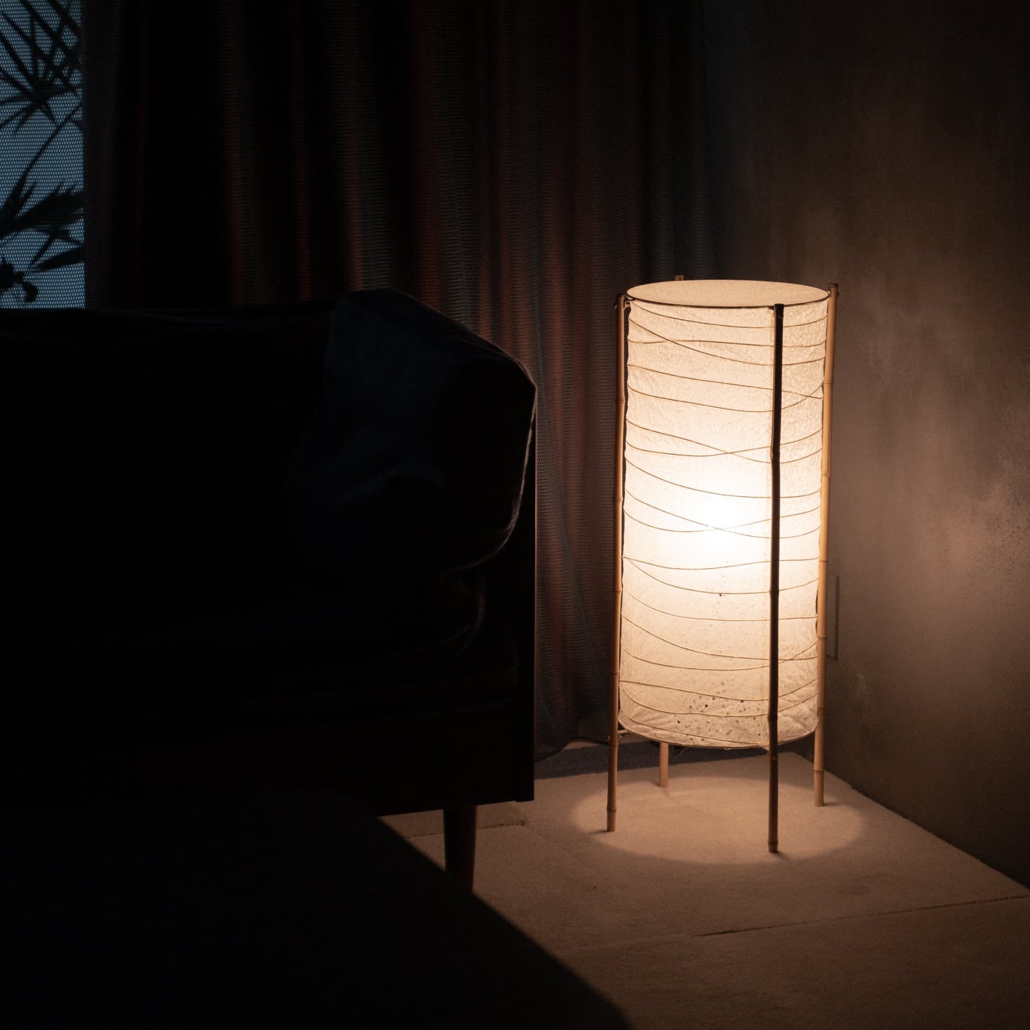 Cylindrical Japanese washi paper lamp with bamboo frame placed in the corner of a dim room, softly illuminating a nearby sofa