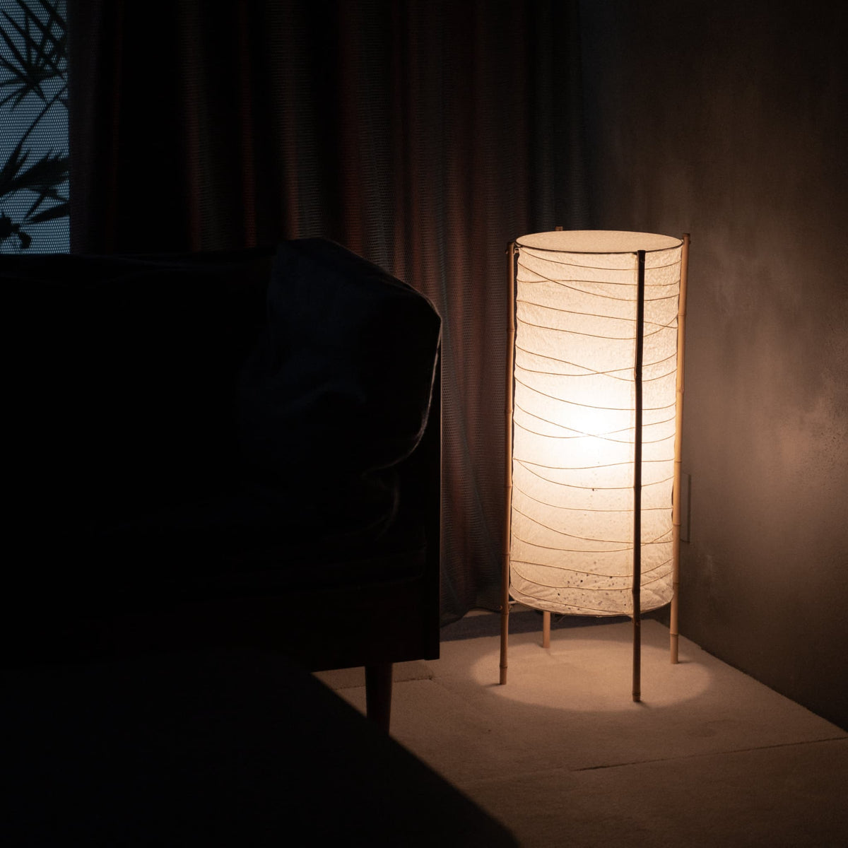 Cylindrical Japanese washi paper floor lamp with bamboo frame placed in the corner of a dim room, softly illuminating a nearby sofa in Japandi living room.