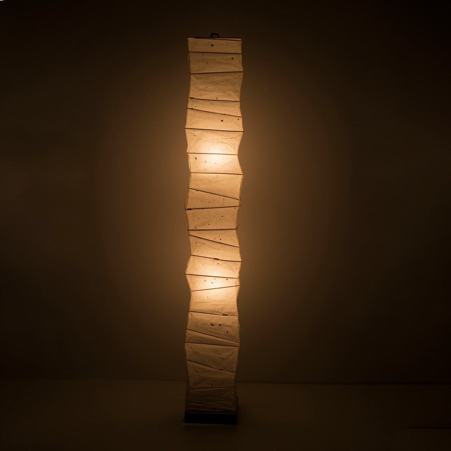 Full front view of a tall, randomly curved Japanese washi paper lamp emitting a warm glow on a dark brown background