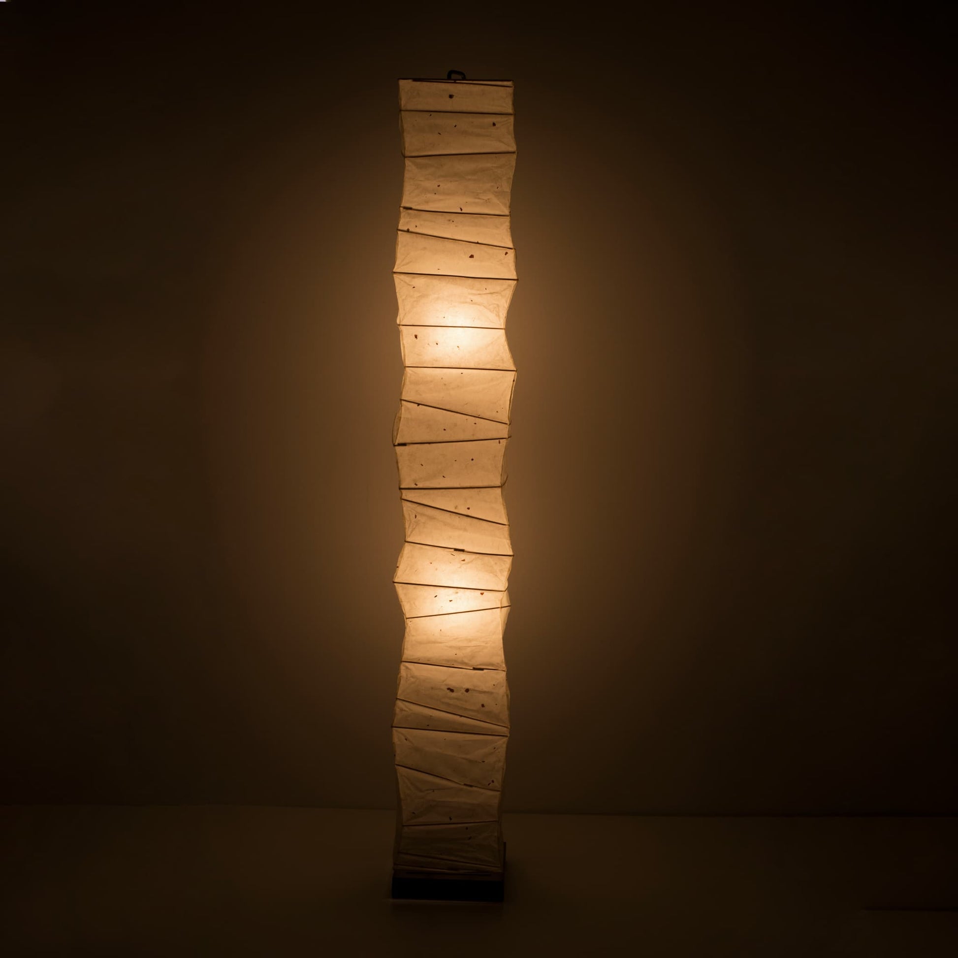 Full front view of a tall, randomly curved Japanese washi paper lamp emitting a warm glow on a dark brown background