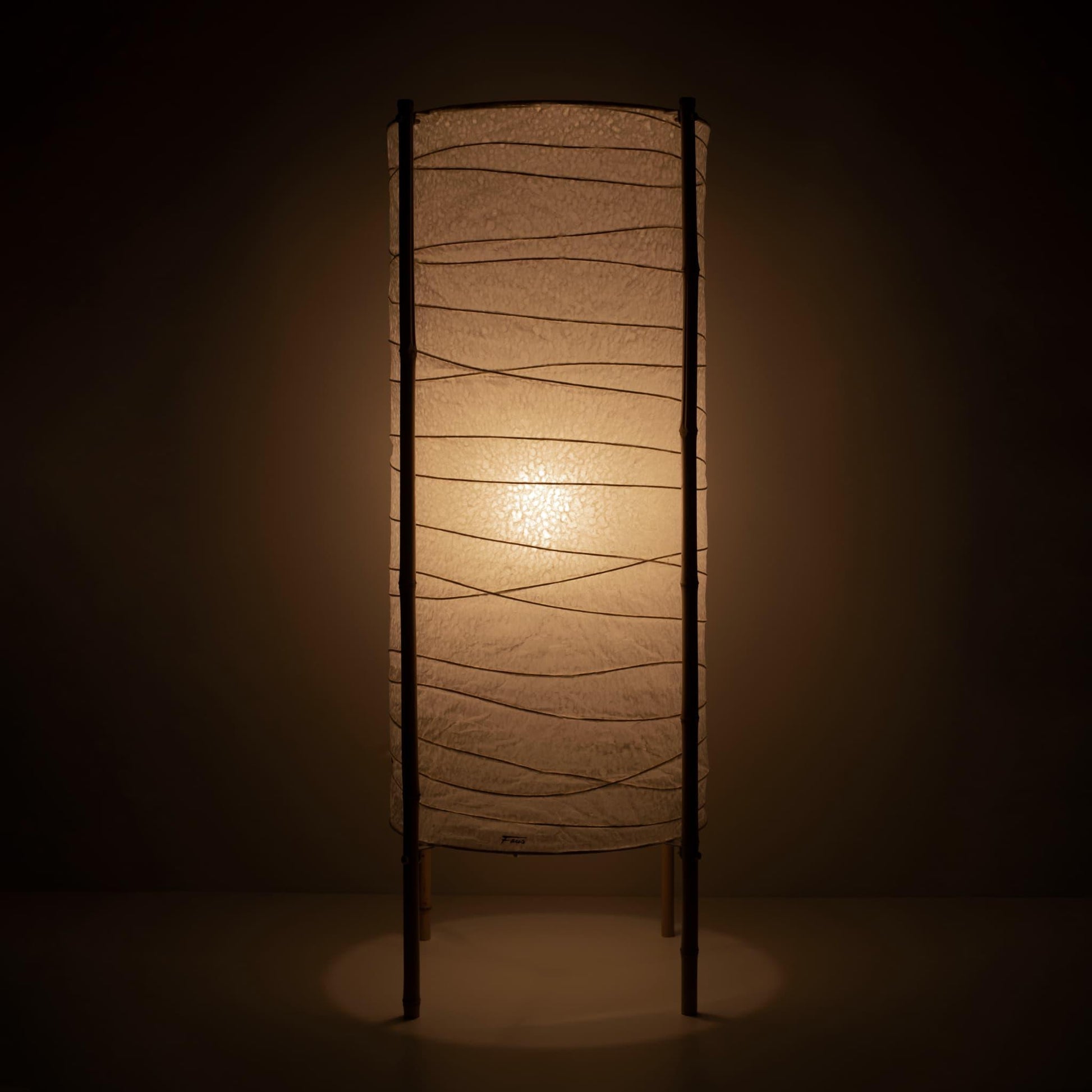 Full view of a cylindrical Japanese lamp with washi paper shade and bamboo frame, warm glow on brown background