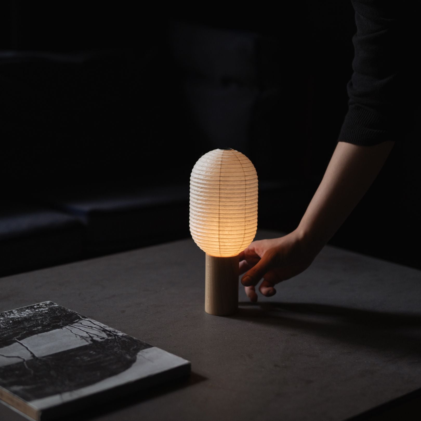 Hand touching a Japanese Paper Table Lamp "TORCHIN VERTICAL" on a gray table in a dimly lit room.