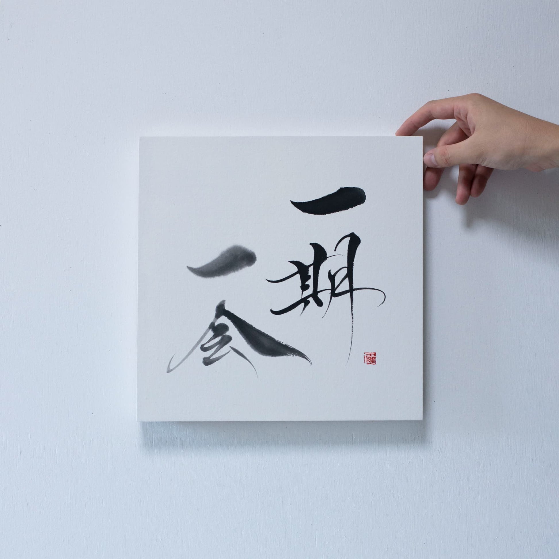 Close-up of a hand holding the corner of a square panel featuring 'Ichigo Ichie' in Japanese calligraphy, mounted on a white wall