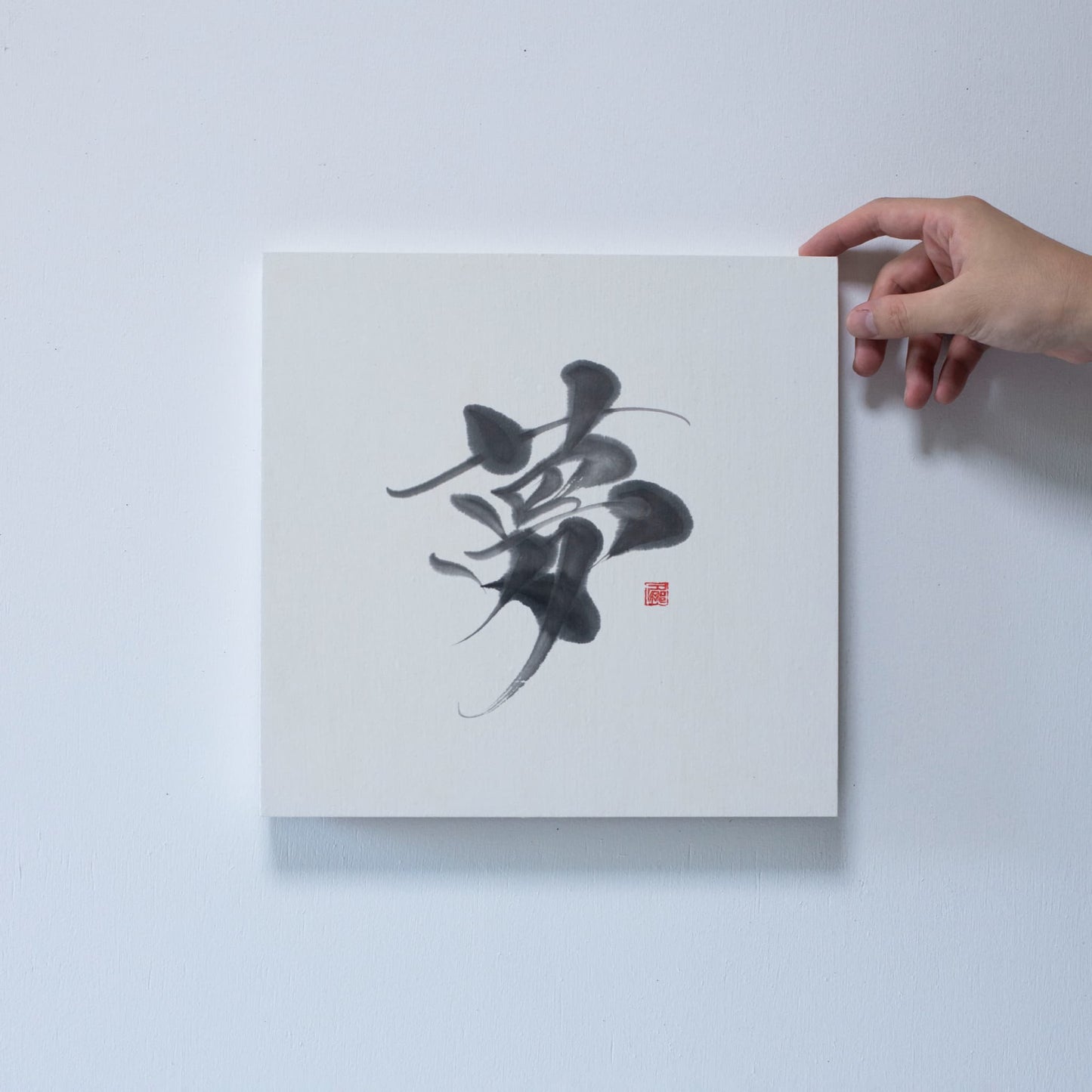 Close-up of a hand holding the corner of a square panel with '夢' (dream) in Japanese calligraphy, displayed on a white wall