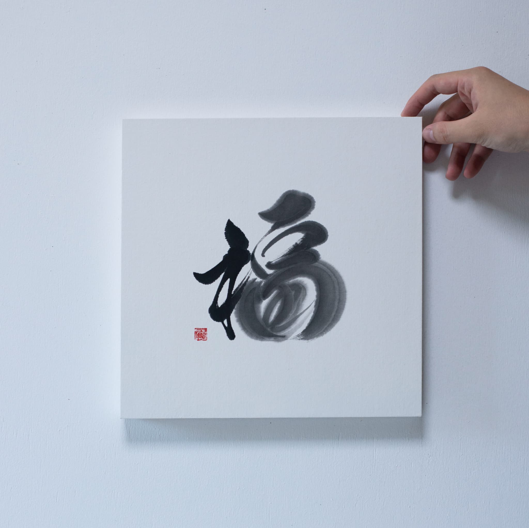 A hand holding the corner of a square panel with Japanese calligraphy of the character '福' (fortune), mounted on a white wall