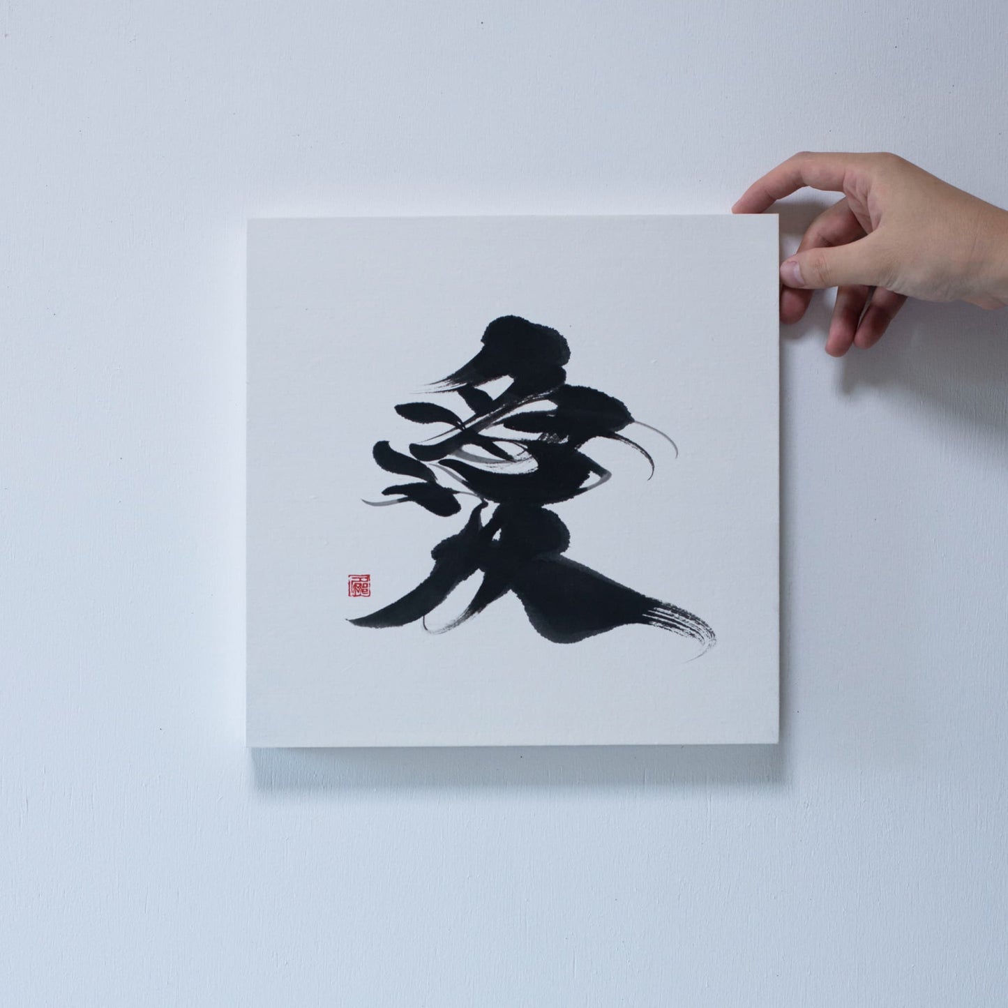 Close-up of a hand holding the corner of a square panel featuring '愛' (love) in Japanese calligraphy, mounted on a white wall