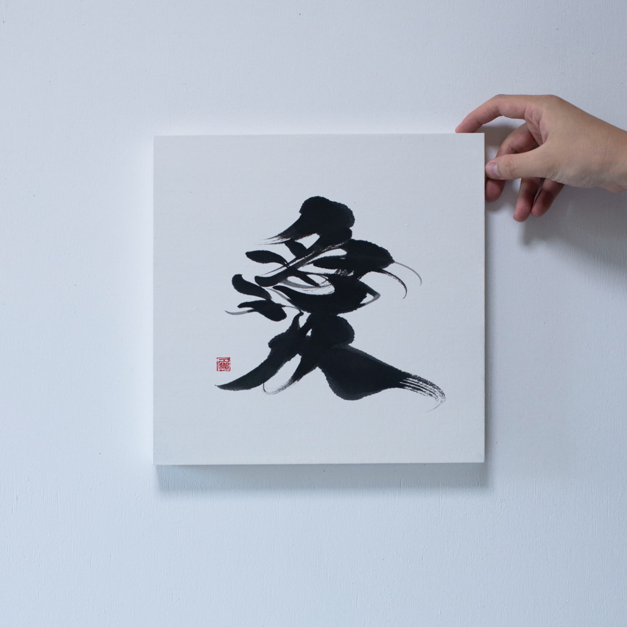 Close-up of a hand holding the corner of a square panel featuring '愛' (love) in Japanese calligraphy, mounted on a white wall