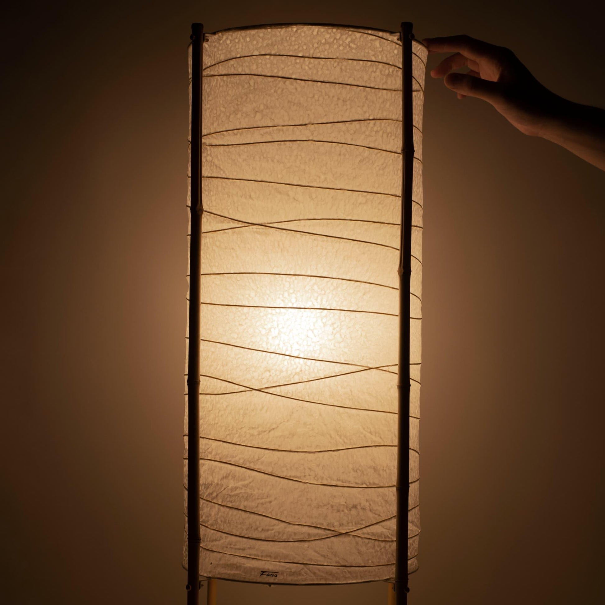 Close-up of a hand touching the top of a cylindrical Japanese washi paper lamp with bamboo frame, warm light on brown background
