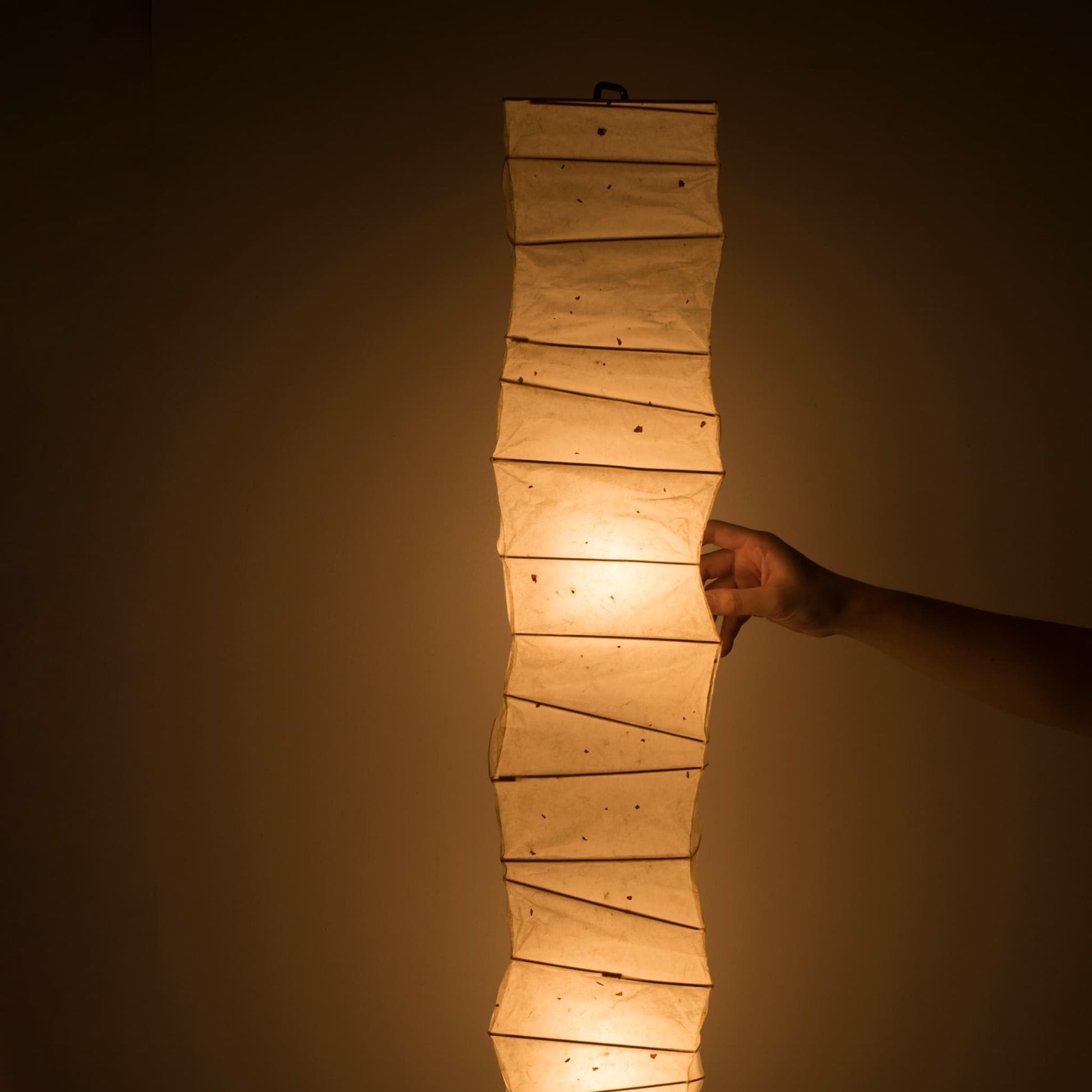 Close-up of a hand touching the side of a curved Japanese washi paper lamp, warm light on brown background