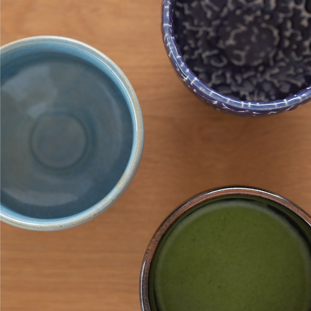 Overhead view of three handcrafted Japanese matcha chawan bowls (blue, dark blue Kairagi, and black) on a wooden table.