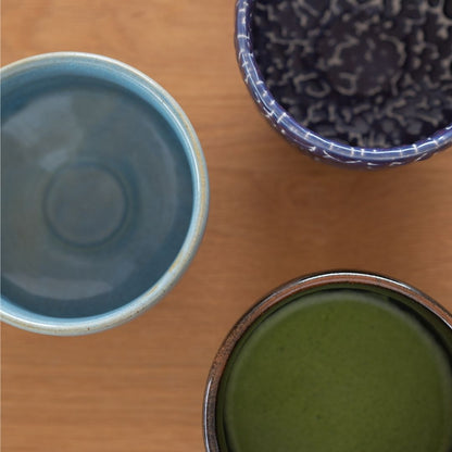 Overhead view of three handcrafted Japanese matcha chawan bowls (blue, dark blue Kairagi, and black) on a wooden table.