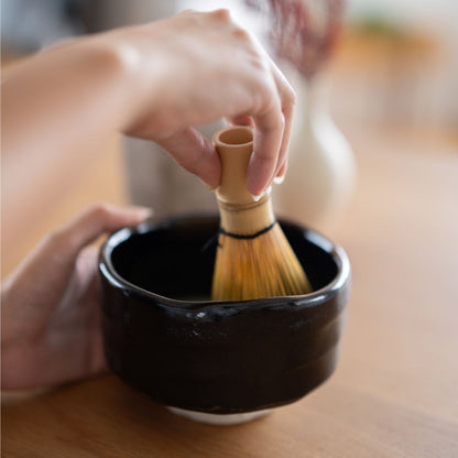 A hand holding a bamboo whisk, preparing matcha in a black handcrafted Mino Ware Japanese bowl.
