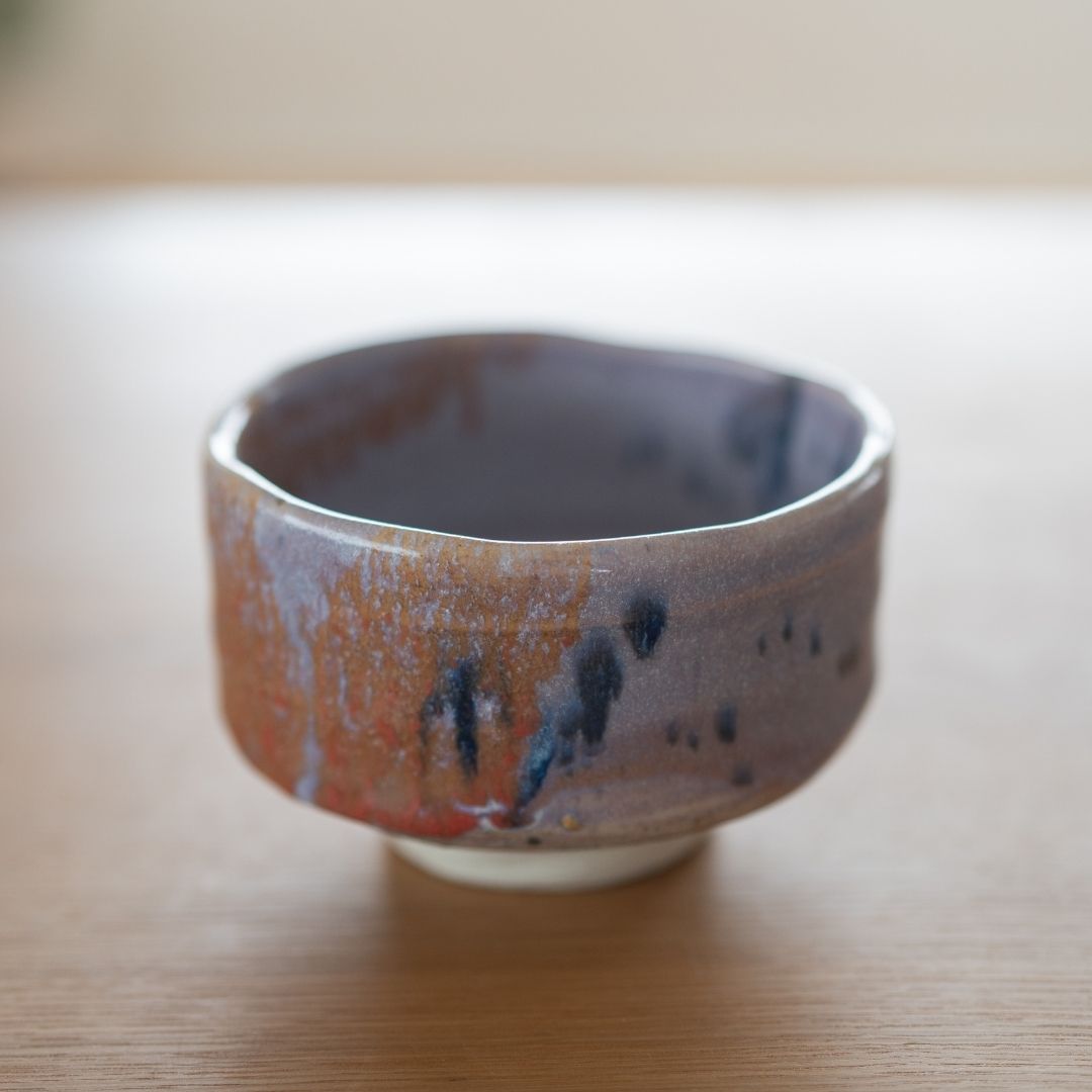 Close-up shot of a single handcrafted Japanese matcha bowl (chawan) with a bold Navy and Red Splash glaze over a Kinyo purple base, resting on a wooden table.