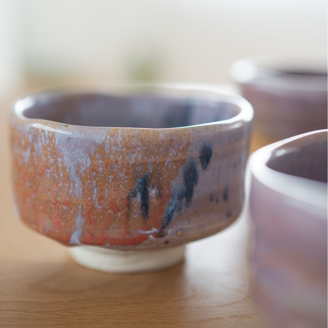 Detailed photo of the Navy and Red Splash Japanese matcha bowl, featuring the abstract glaze pattern and white foot. Partially blurred view of two other chawan bowls suggests a collection or set.