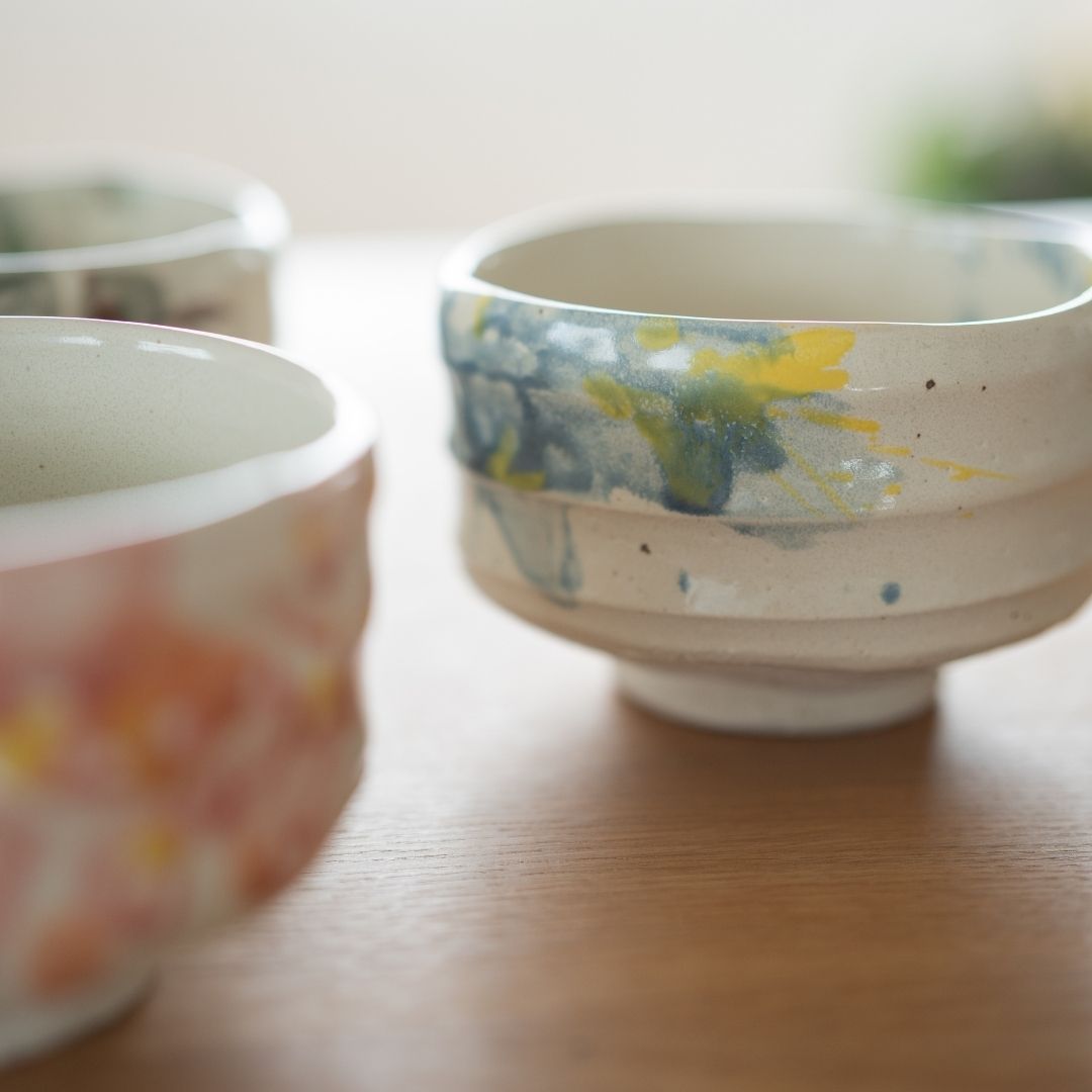 Focus on the "Splash" Japanese matcha bowl with blue and yellow glazes, displayed on a wooden table alongside the pink "Flower" bowl, showcasing the variety of handcrafted Shino ware.