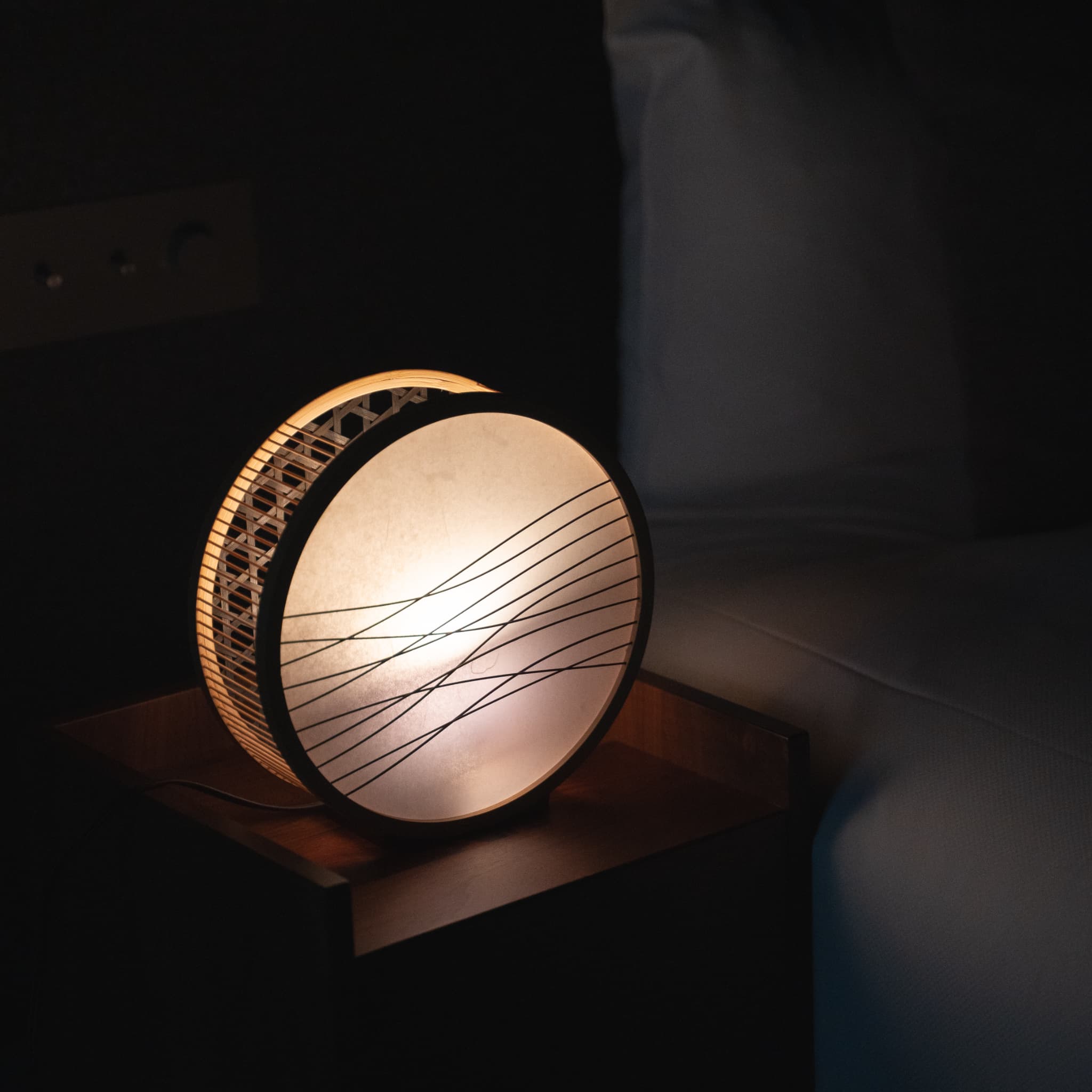 A Japanese Bamboo Floor Lamp “Lantern of Shadow" glowing on a bedside table in a dimly lit bedroom.