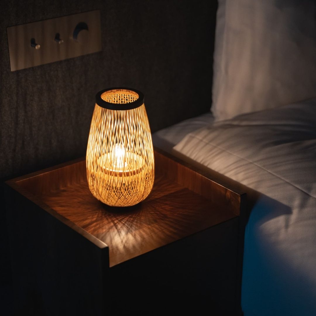 A Japanese bamboo lamp "Flower bud," handcrafted with traditional bamboo weaving techniques, sits on a wooden bedside table, emitting a warm glow