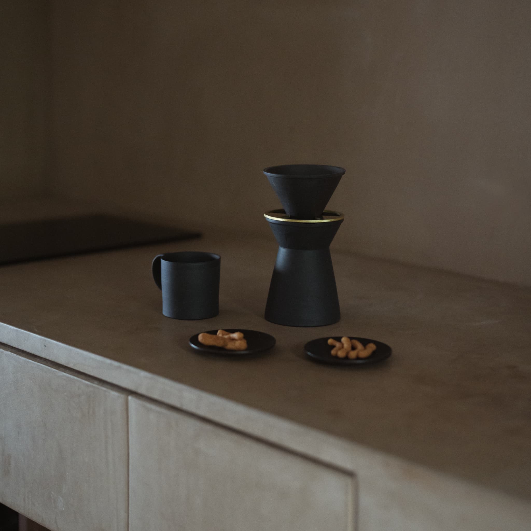 Black Banko-yaki coffee server, dripper, and cup, accompanied by a wagashi sweet on a small black plate, in a beige Japandi-style kitchen.