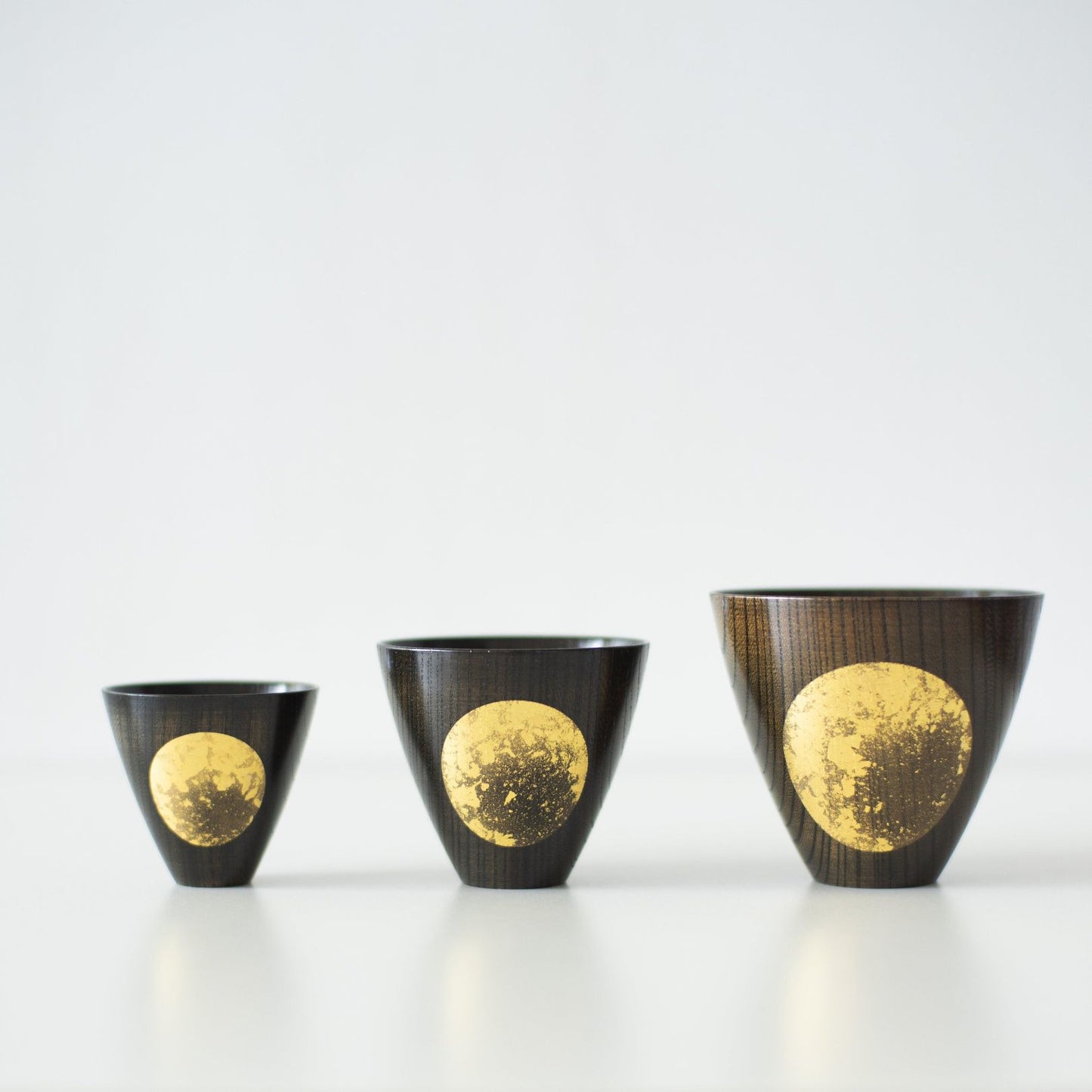 Three Japanese lacquerware sake cups, each adorned with a moon design in gold leaf, are lined up.