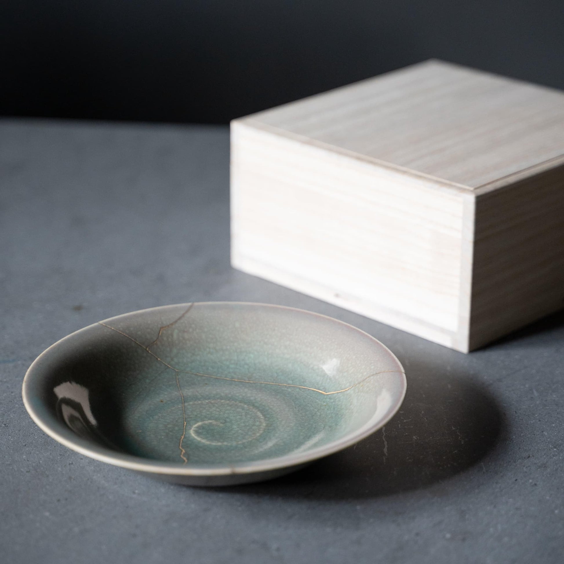 Pale green bowl repaired with Kintsugi, a traditional Japanese technique, presented in a paulownia wood box.