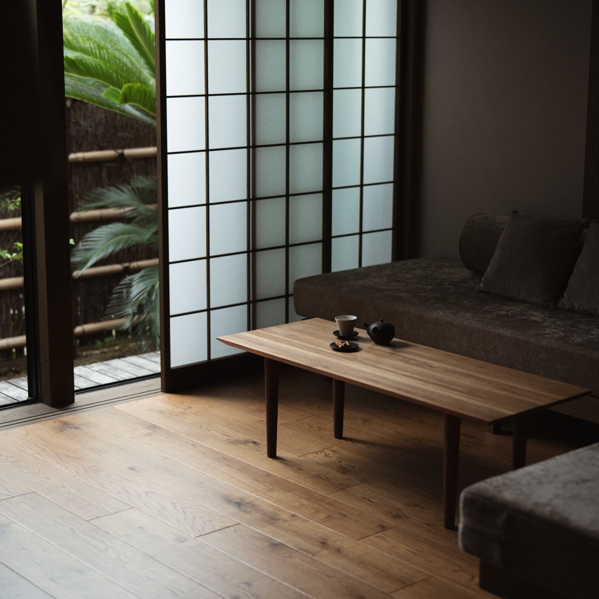 Japandi living room with low sofa and wooden table, bathed in natural light filtering through shoji screens revealing lush greenery outside.