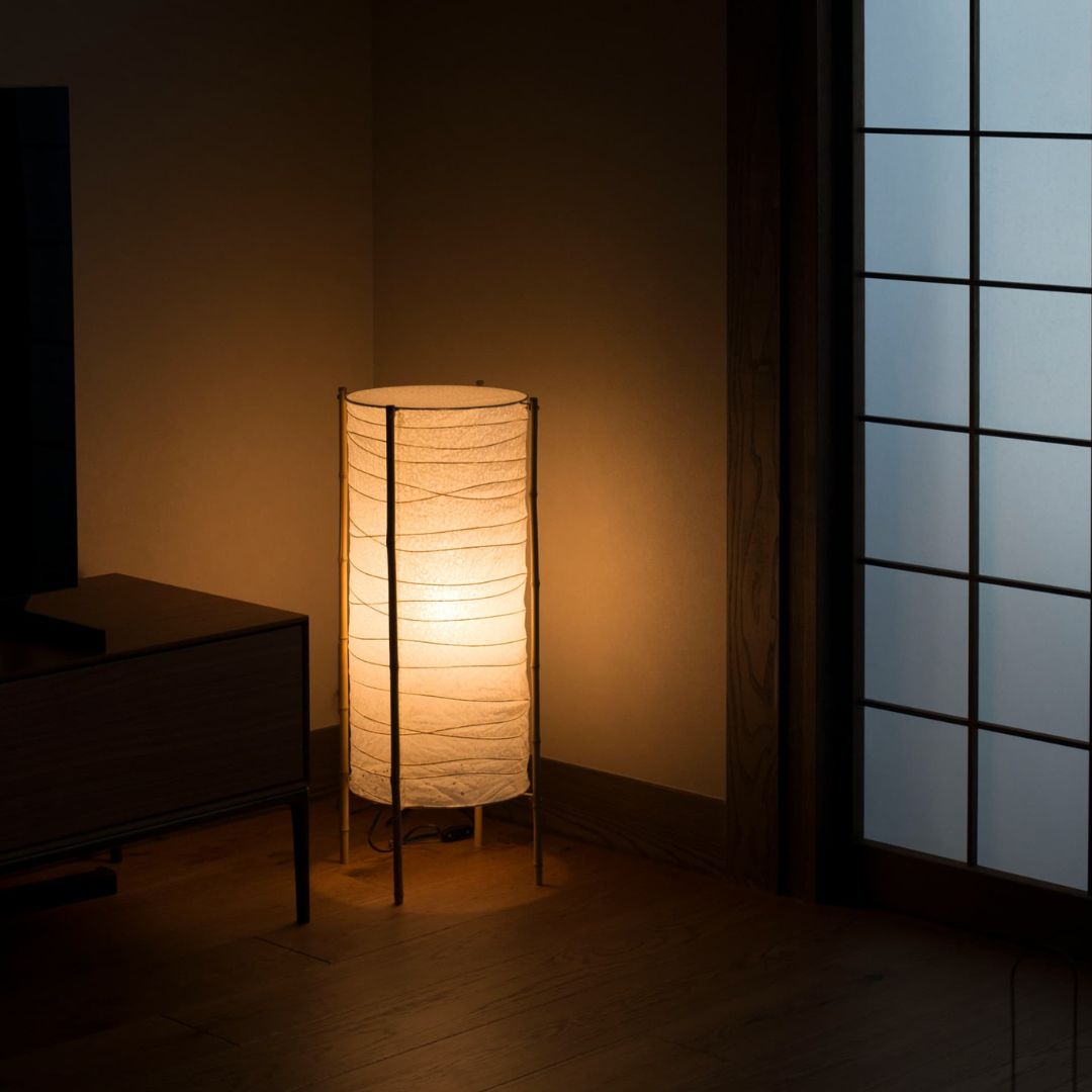 A cylindrical Japanese Paper Floor Lamp "NiPPON" emits a warm light in the corner of a dimly lit room. A black sofa is positioned to its left.