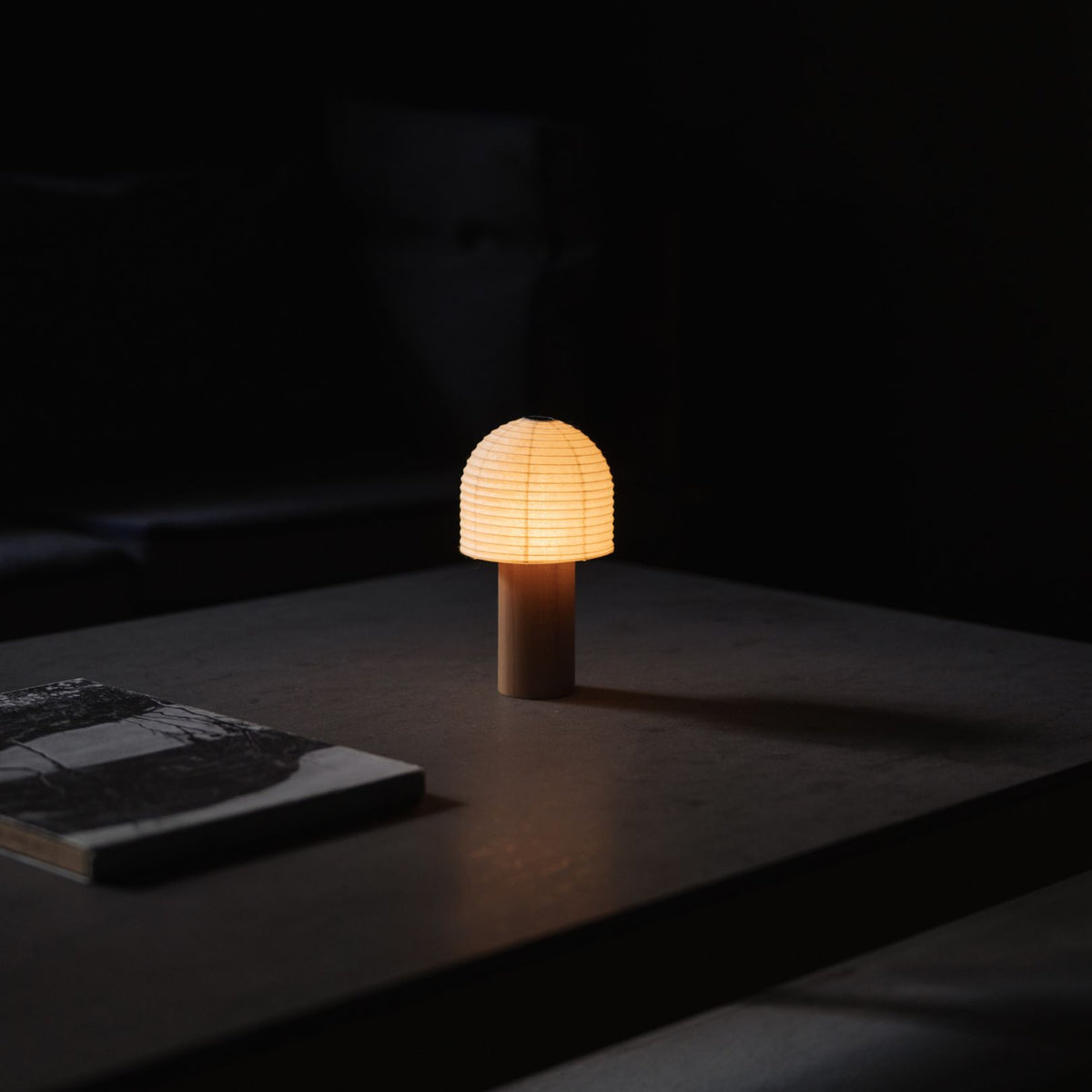 Japanese Paper Table Lamp "TORCHIN BELL" emitting warm light on a gray table in a dimly lit room.