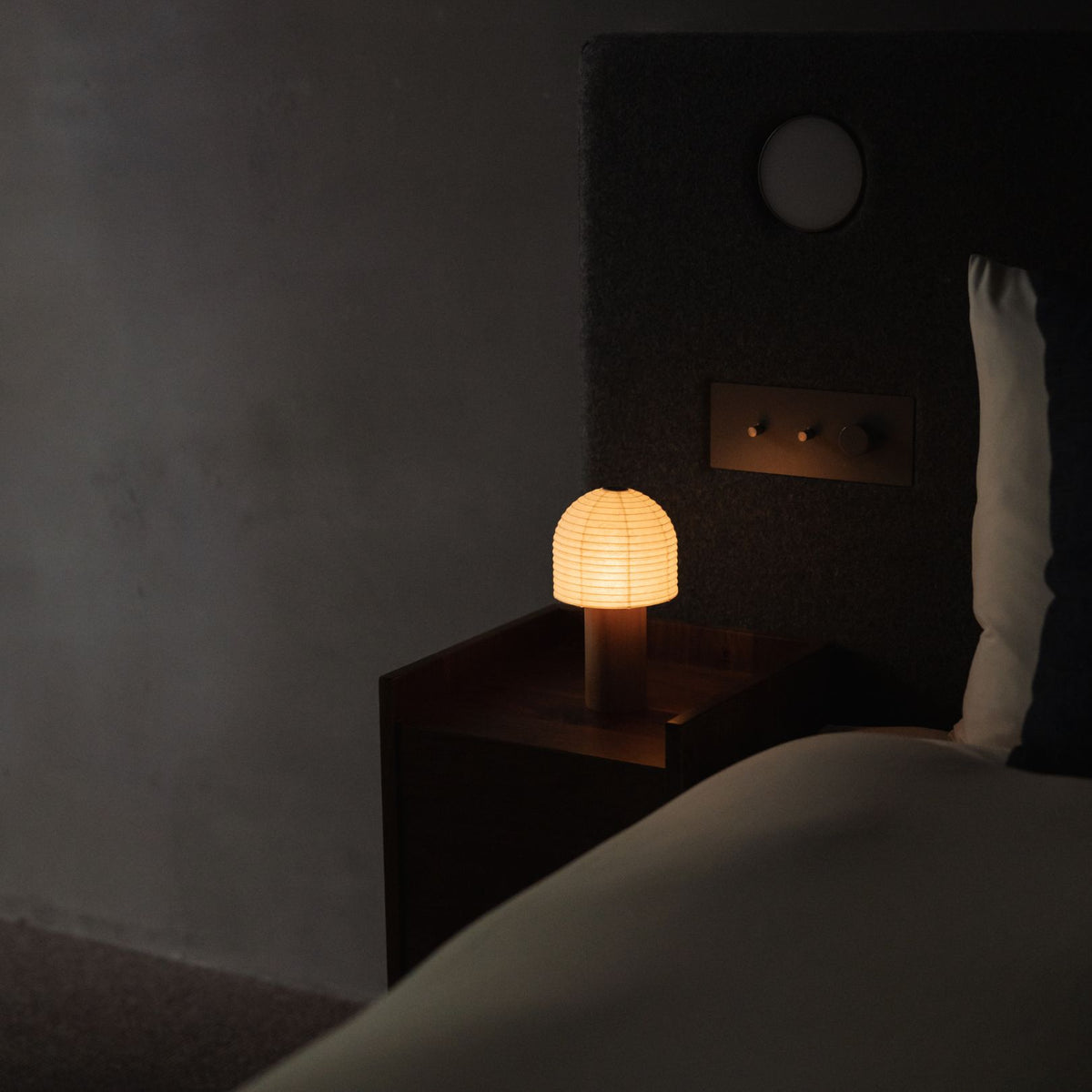 Side view of a Japanese Paper Table Lamp "TORCHIN BELL" emitting warm light on a bedside table in a Japandi bedroom