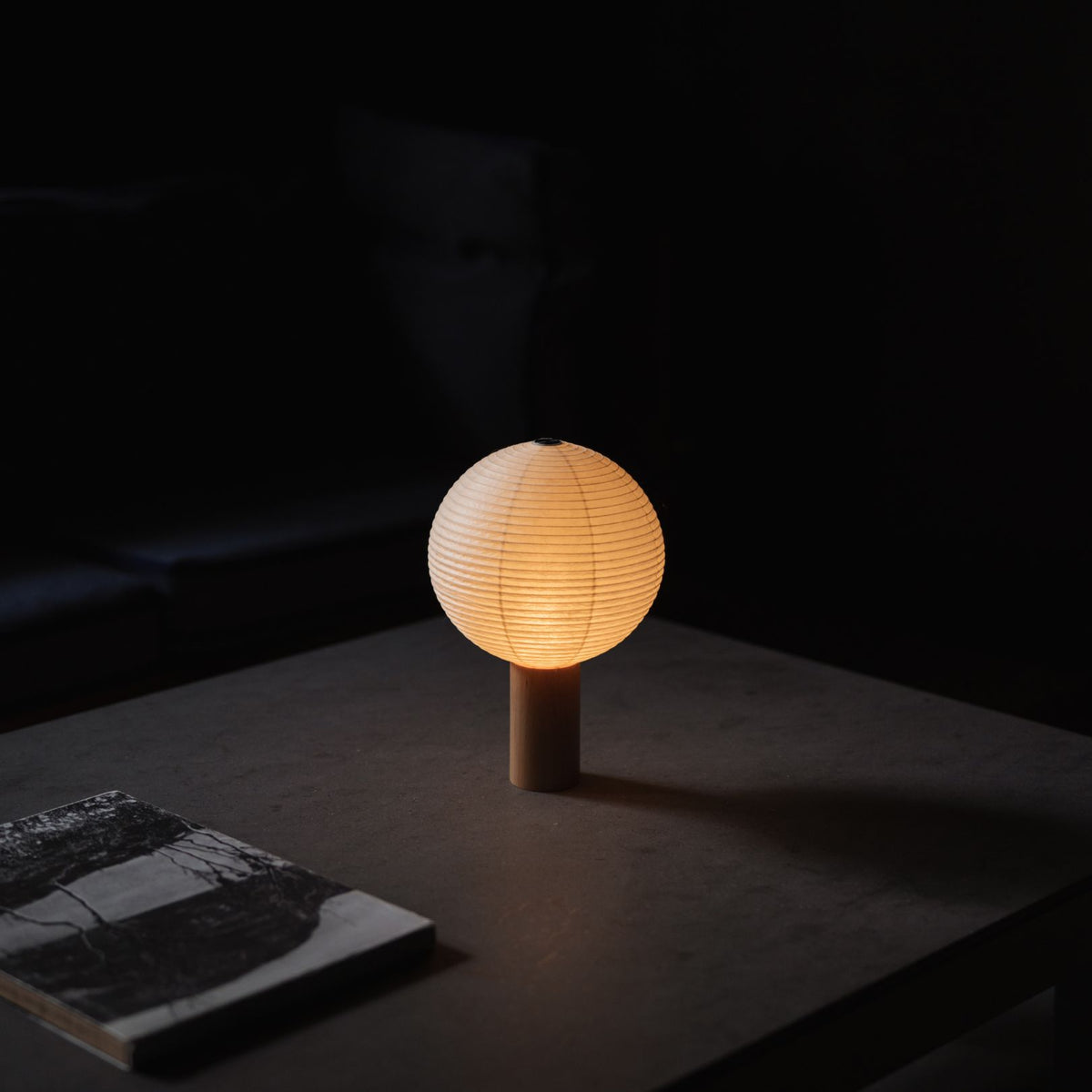 Japanese Paper Table Lamp "TORCHIN CIRCLE" emitting warm light on a gray table in a dimly lit room.