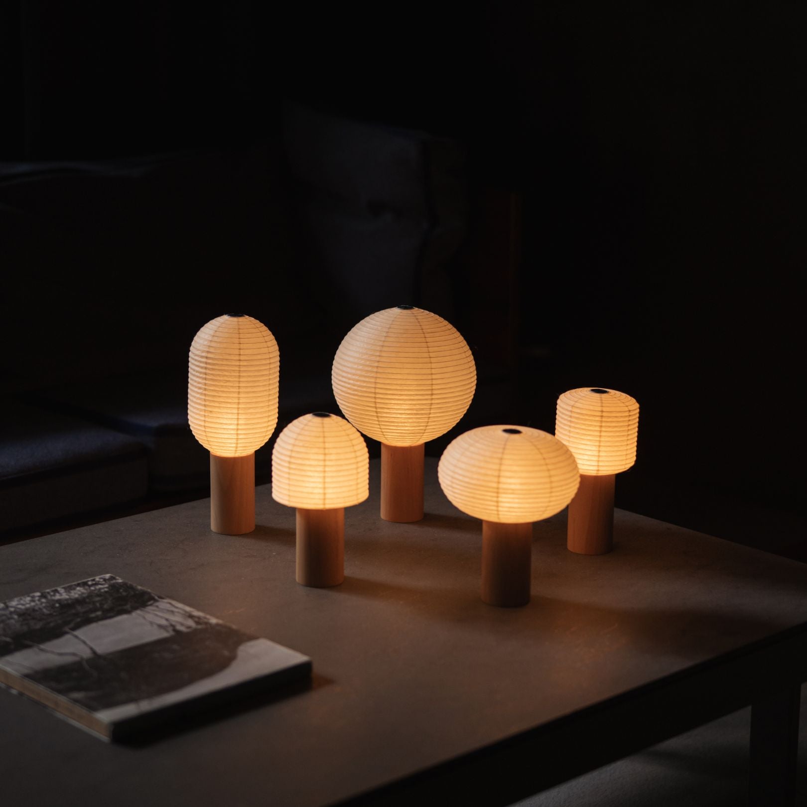 Five different Japanese Paper Table Lamps "TORCHIN" emitting warm light on a gray table in a dimly lit room.