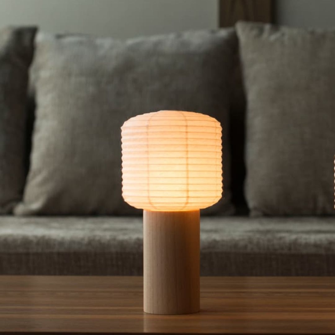 A side-view photo of a Japanese paper portable lamp, "Torchin," placed on a wooden table. A gray sofa is in the background. The Torchin's lampshade is made from a Japanese lantern with a cylindrical wooden handle attached beneath.