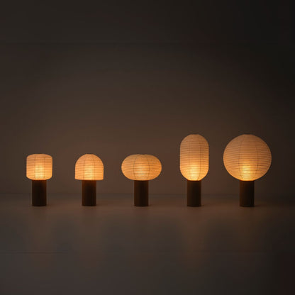 Five portable lamps, "TORCHIN", with warm lamp, lined up in a dimly lit room. The lampshades are made of Japanese washi paper from Yame-chochin lanterns, and each lamp has a wooden cylindrical handle.