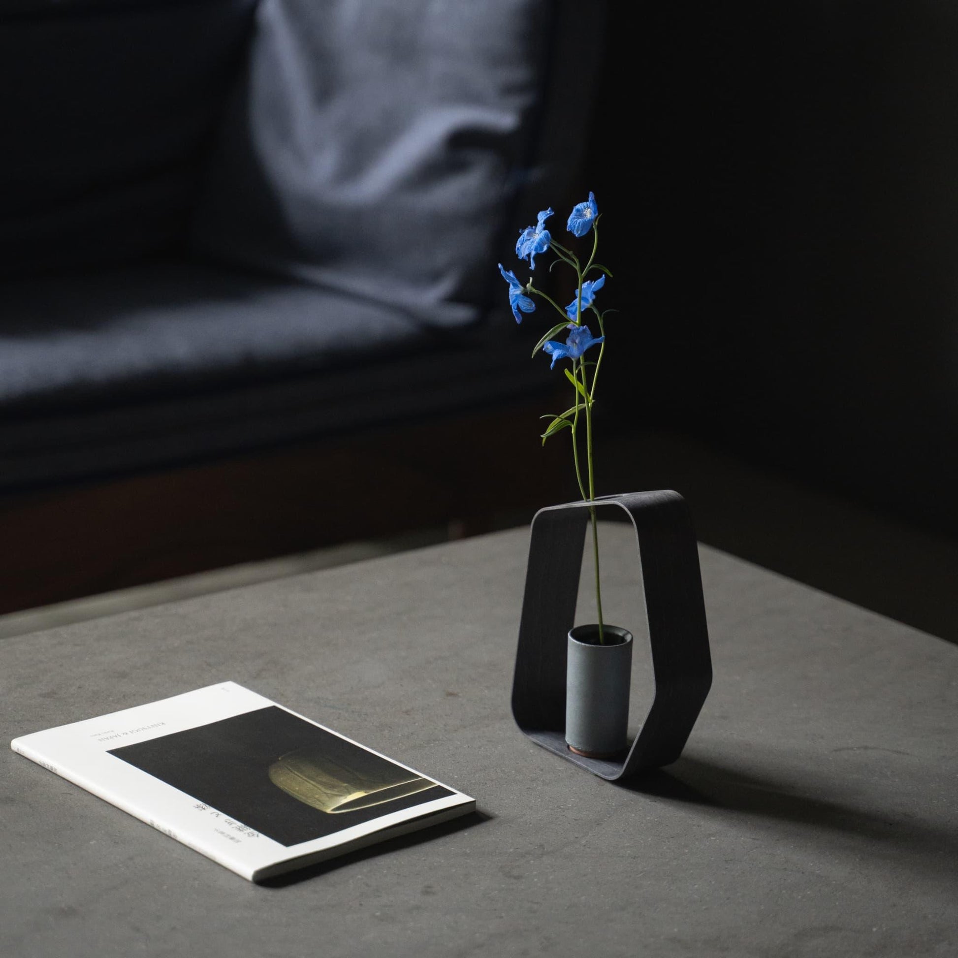 A minimalist black iron vase crafted using traditional Japanese roof tile making techniques, paired with a black wood frame, placed on a grey Japandi style table.