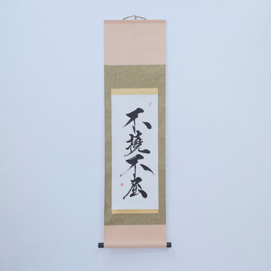 A brown traditional Japanese hanging scroll with calligraphy of '不撓不屈' (Indomitable Spirit) written on rice paper, displayed on a white wall