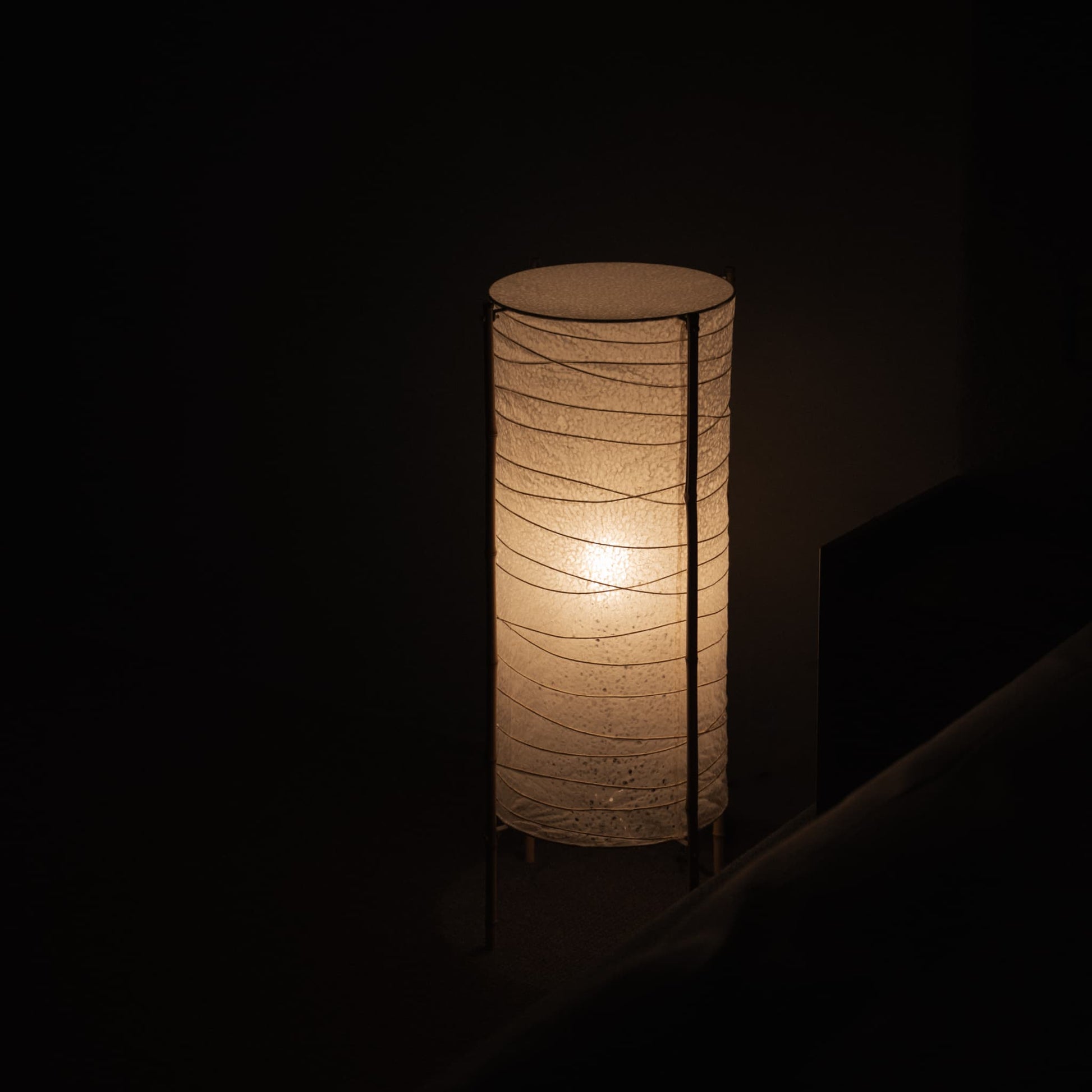 Dimly lit room with a Japanese cylindrical washi paper lamp and bamboo frame, casting a warm glow