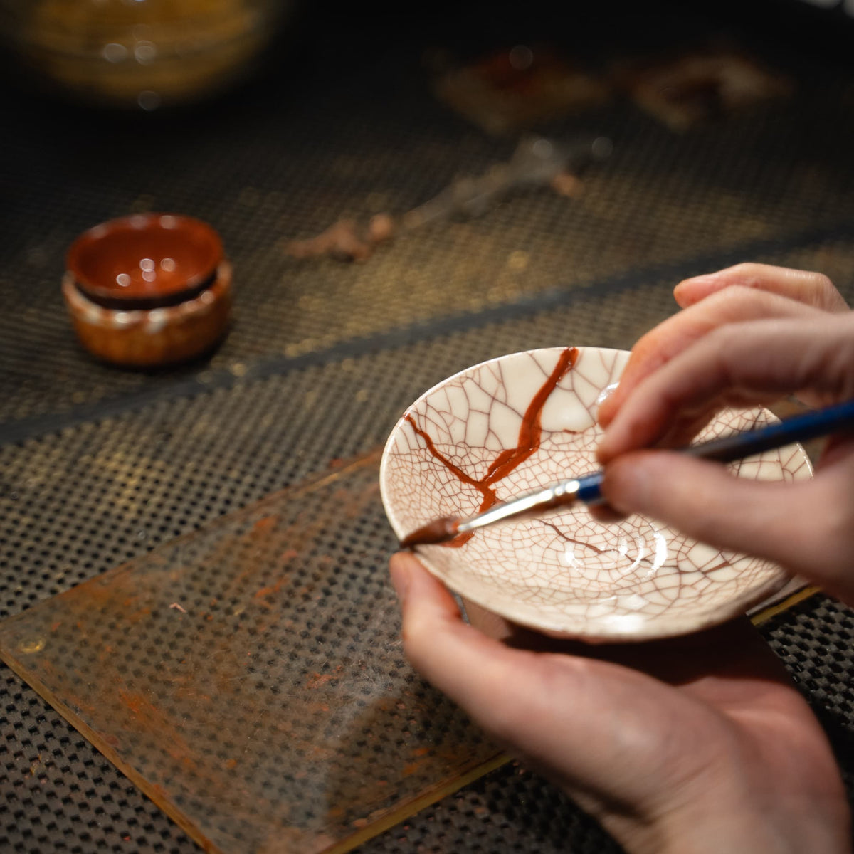 An artisan's hands apply Urushi lacquer to bond a broken white Kintsugi bowl.