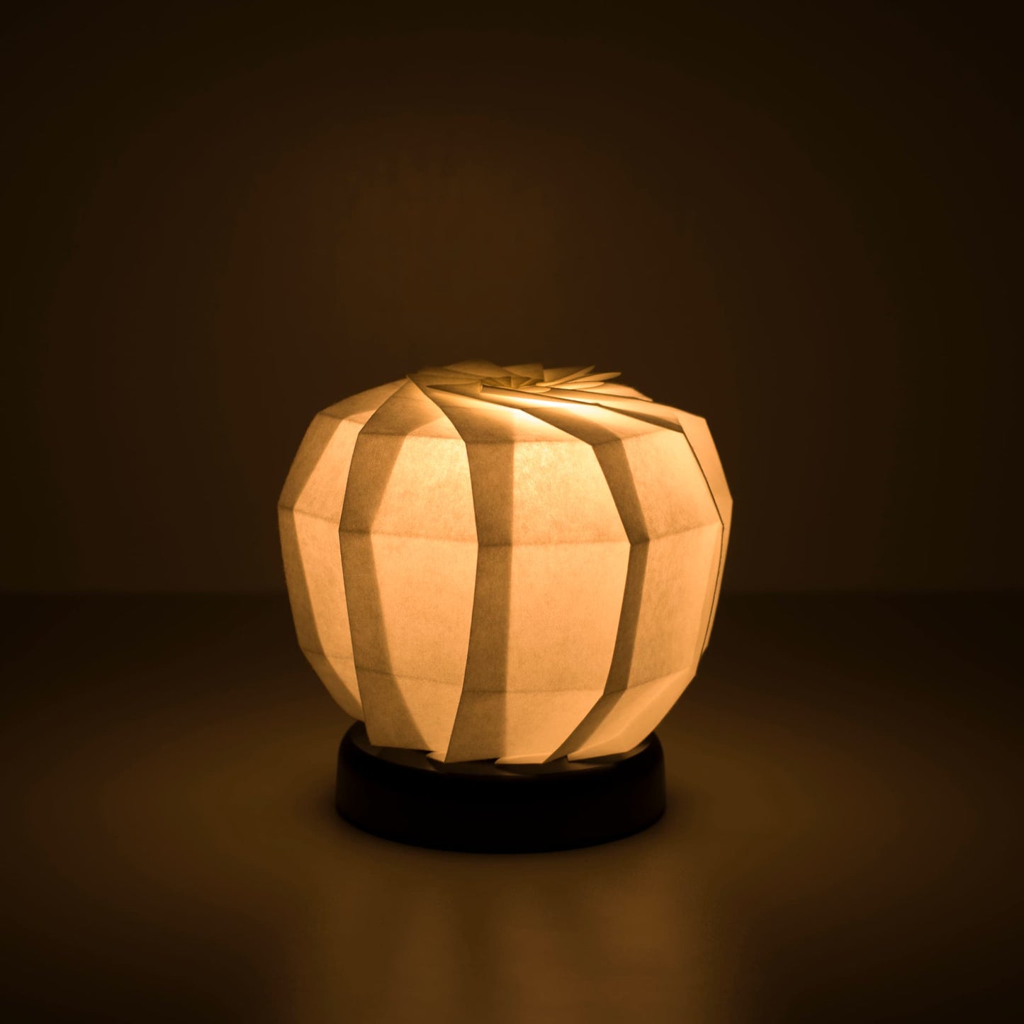 Origami Table lamp "maru M" with a black base and a spherical white lampshade crafted from intricately folded washi paper,  emitting a warm and inviting light.